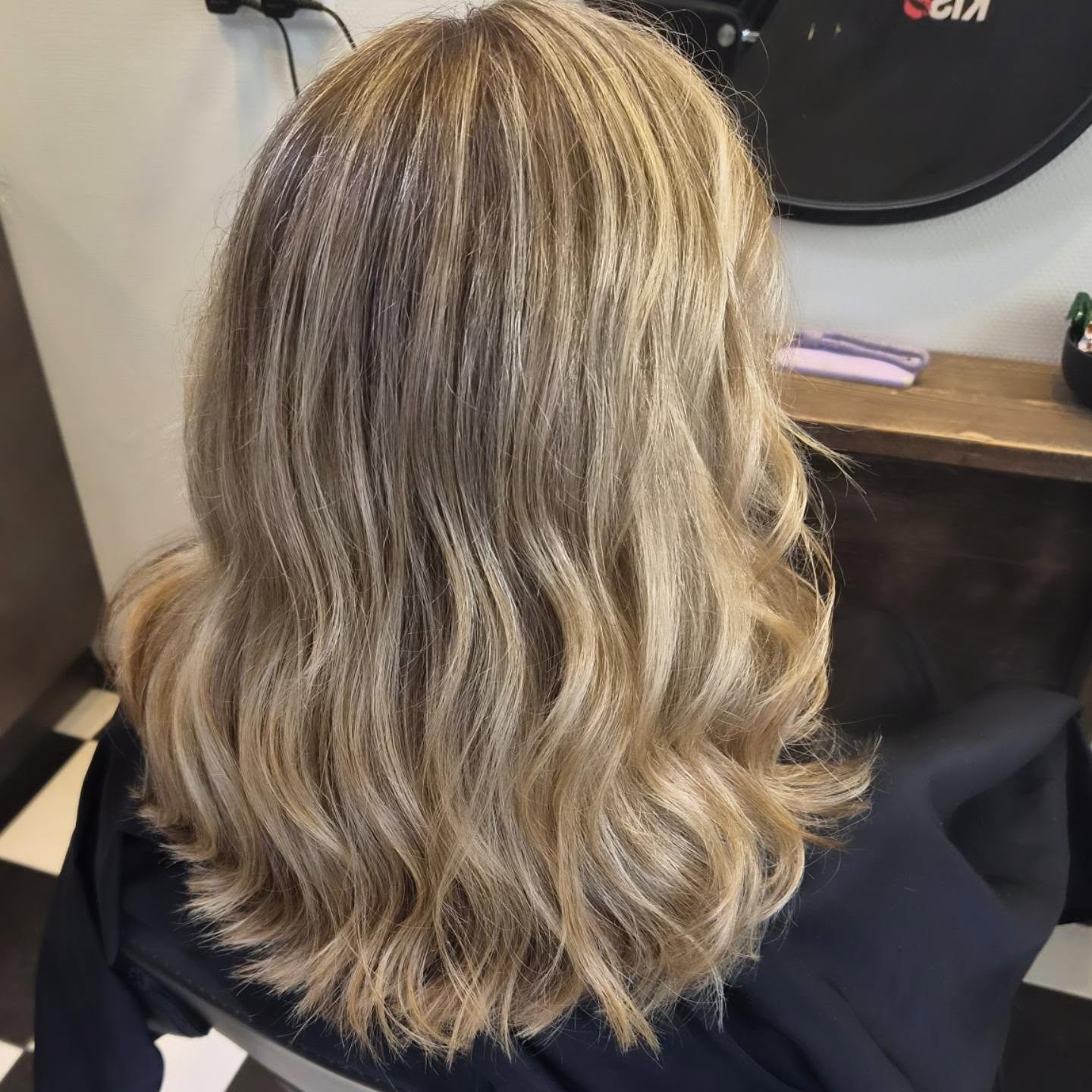 Halflang blond haar met babylights en zachte golven, van achteren gezien in de salon